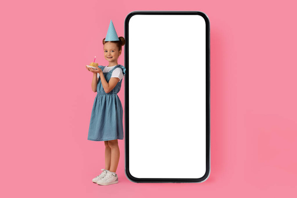 Roztomilé holčička s narozeninovým dortem stojící v blízkosti Big Blank Smartphone s bílou obrazovkou, Happy veselý předpubertální ženské dítě slaví B-Day a doporučuje nové mobilní aplikace, Mockup - Fotografie, Obrázek