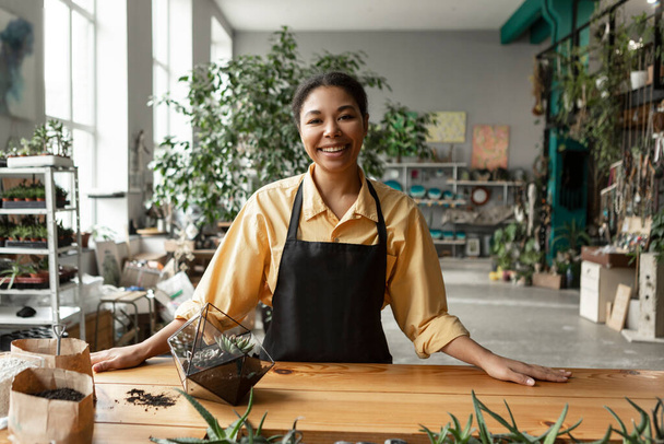 Šťastný africký americký zahradník pracující ve skleníku, vyrábějící florária, stojící u stolu ve skleníku, vytvářející uměleckou botanickou kompozici v teráriu - Fotografie, Obrázek