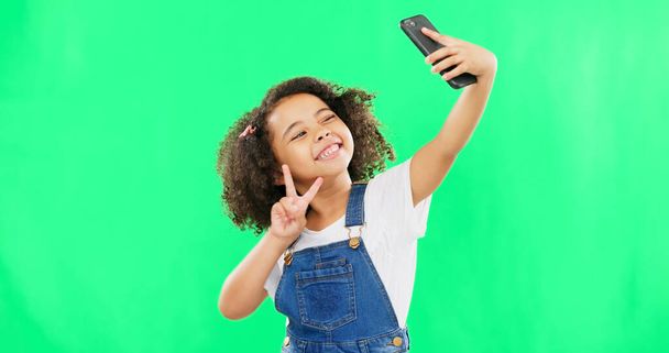 プロフィール写真から幸せとスタジオで電話、自撮り、子供の平和のサイン。孤立した、緑の背景と笑顔でソーシャルメディアのためのv手ジェスチャーを示す技術を持つ若い女の子. - 写真・画像