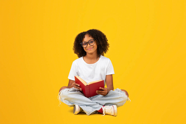 Cheerful smart curly teenager black schoolgirl in white t-shirt and glasses reads book, isolated on yellow background, studio. Study, knowledge, hobbies, education and homework, exam preparation - Fotó, kép