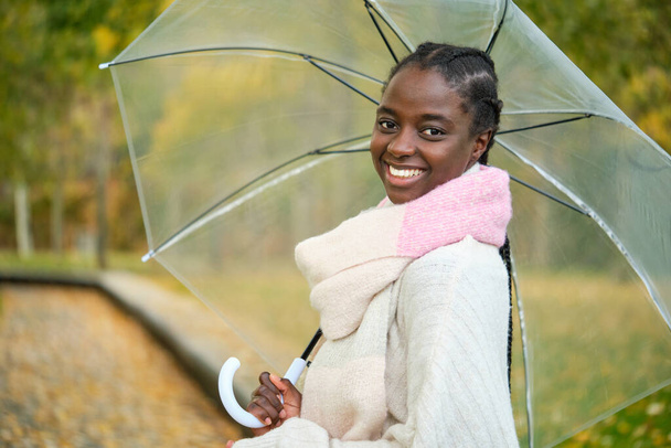 Портрет африканки с прозрачным зонтиком, смотрящей в камеру и улыбающейся осенью. - Фото, изображение