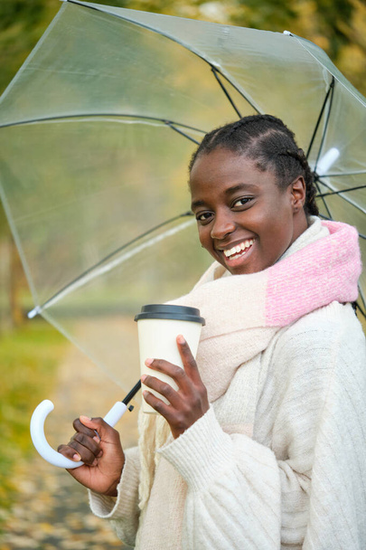 Donna africana con un caffè e ombrello trasparente guardando la fotocamera e sorridendo in autunno. - Foto, immagini