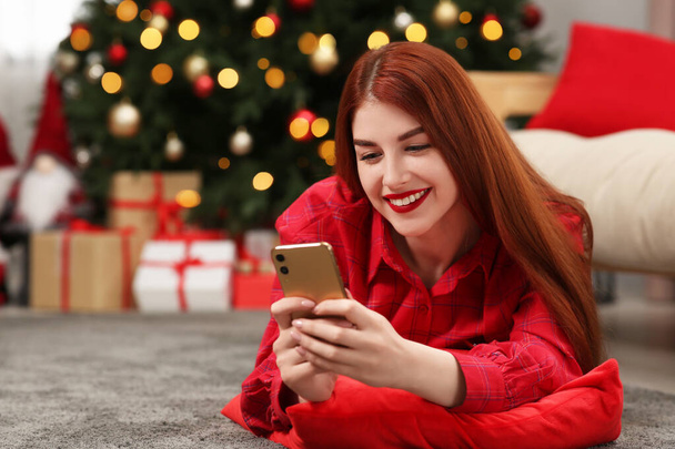 Hermosa mujer joven utilizando el teléfono inteligente cerca del árbol de Navidad en casa, espacio para el texto - Foto, imagen
