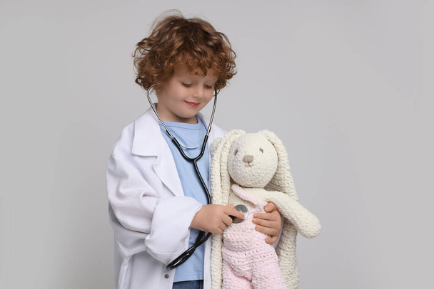 Мальчик играет в доктора с игрушечным кроликом на светло-сером фоне - Фото, изображение