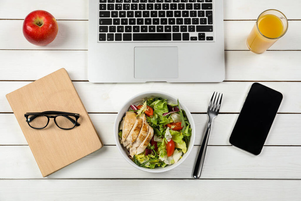 Bowl with tasty food, glass of juice, laptop, apple, book and smartphone on white wooden table, flat lay. Business lunch - Photo, Image