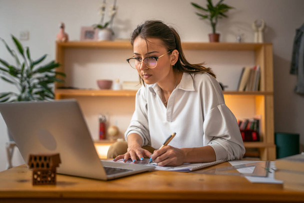 nuori valkoihoinen nainen opiskelee kannettavan tietokoneen ääressä kotona - naispuolinen lukukirja valmistelee tenttiä tai toimeksiantotestiä, jossa on online-konsultointi - koulutuksen ja oppimisen käsite - Valokuva, kuva