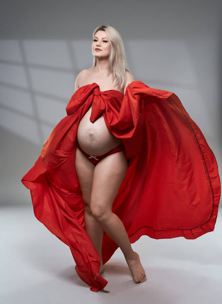 Beautiful pregnant young blonde woman in red wavy sheet on gray background - Foto, Bild