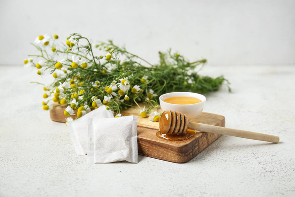 Wooden board with bowl of honey, tea bags and fresh chamomile flowers on light background - Photo, Image