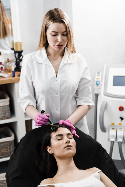 Proces masażu twarzy z czarnym kamiennym wałkiem. Kosmetolog używający kamiennego wałka do masażu twarzy dla pacjentki. Masaż twarzy wałkiem kosmetycznym w klinice kosmetologii - Zdjęcie, obraz