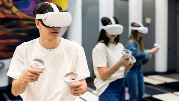 Ryhmä kolme nuorta ystävää pelaa VR pelejä areenalla käyttäen ohjaimet ja kuulokkeet - Valokuva, kuva
