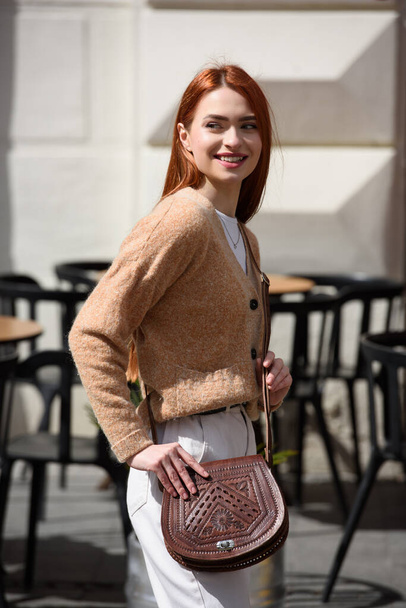een roodharig meisje in witte broek en een beige trui poseert buiten met een kleine leren handtas. - Foto, afbeelding