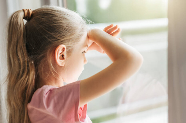 Одинокая маленькая девочка смотрит в окно, стоя дома одна.  - Фото, изображение