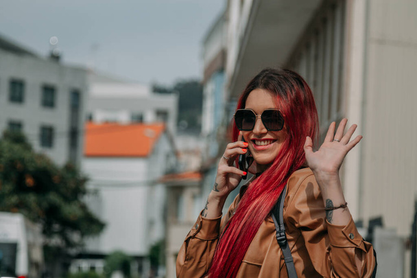 happy young woman with red hair waving and talking on the phone in the street - Foto, Imagen