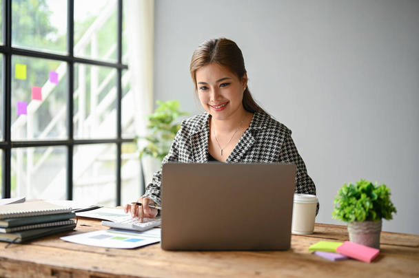 Piękna milenijna azjatycka bizneswoman siedzi przy biurku w swoim biurze i skupia się na swojej pracy na laptopie. - Zdjęcie, obraz