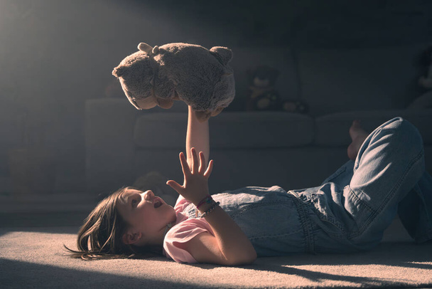 Το παιδί παίζει και μιλάει σε βελούδινο παιχνίδι. Κοριτσάκι που κρατά αρκουδάκι με φως το ηλιοβασίλεμα στο σπίτι. - Φωτογραφία, εικόνα