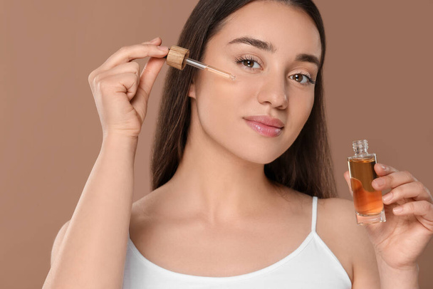 Beautiful woman applying essential oil onto face on brown background - Фото, зображення