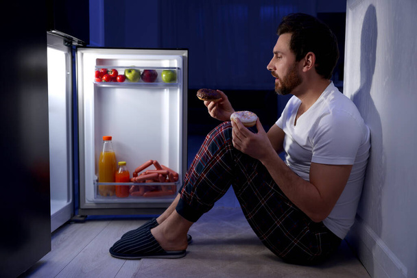 Man with donuts near refrigerator in kitchen at night. Bad habit - Photo, image