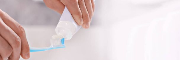 Man squeezing toothpaste from tube onto toothbrush in bathroom, closeup. Banner design with space for text - 写真・画像