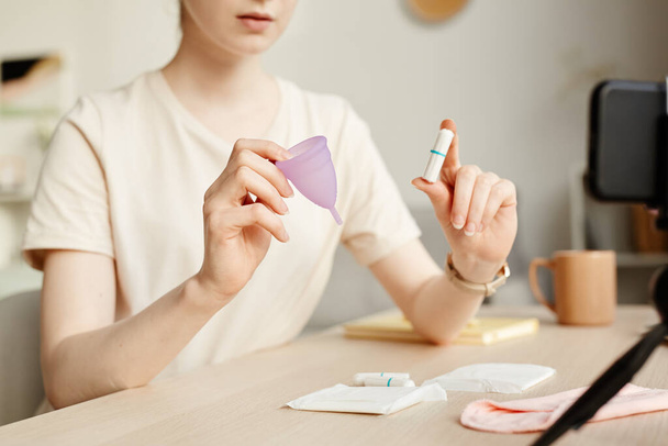 Close up of young woman comparing tampons and menstrual cup on camera and filming educational video, copy space - Photo, Image