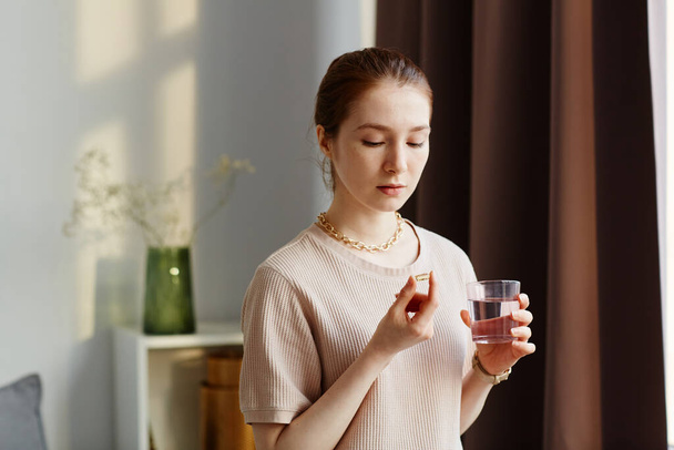 Minimale portret van jonge vrouw met vitamine capsule en het nemen van supplementen met glas water thuis - Foto, afbeelding