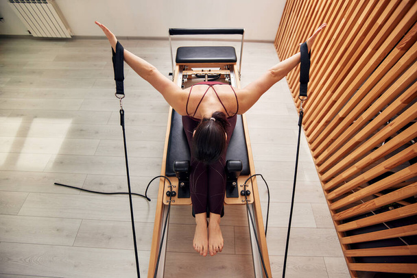 Een jong meisje doet Pilates op een hervormer bed in een heldere studio. Een slanke brunette doet oefeningen om de spieren van haar armen en schoudergordel te versterken. Gezond levensstijl concept. Bovenaanzicht - Foto, afbeelding