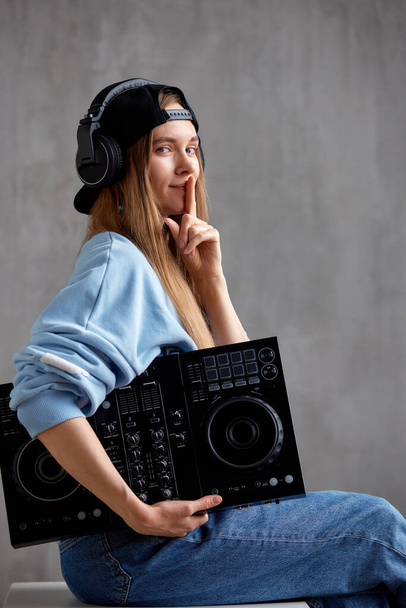 Une jeune DJ aux cheveux longs avec un pull bleu, un jean et une casquette noire pose alors qu'elle est assise avec une console de mixage noire à la main. Plan studio, fond gris - Photo, image