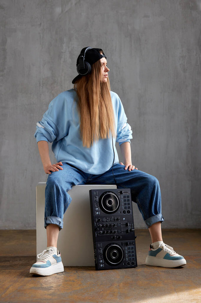 A young pretty long haired DJ girl in a blue sweater, jeans and a black baseball cap is posing while sitting on a white cube. A black DJ mixing console stands nearby. Studio shot, gray background - Photo, Image