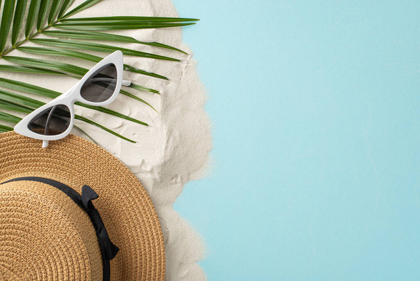 Una vista dall'alto del fine settimana assolato. Attrezzature da spiaggia essenziali come occhiali da sole, cappello da sole e una foglia di palma posizionata contro un ambiente sabbioso e blu pastello, con uno spazio vuoto per testo o annunci - Foto, immagini