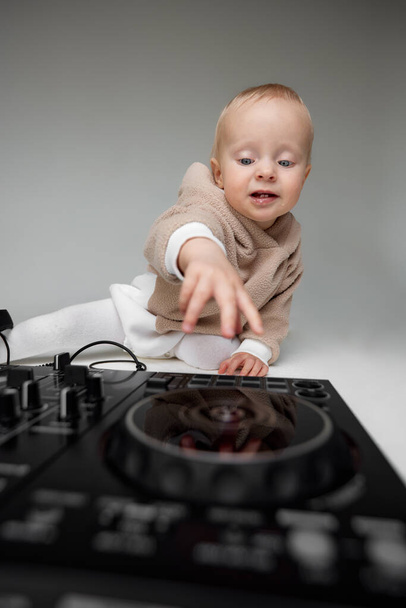 A cute smiling little child in a beige hoodie is sitting on the floor with dj headphones and a dj mixing board. Music and fun. Isolated on grey background - Photo, image