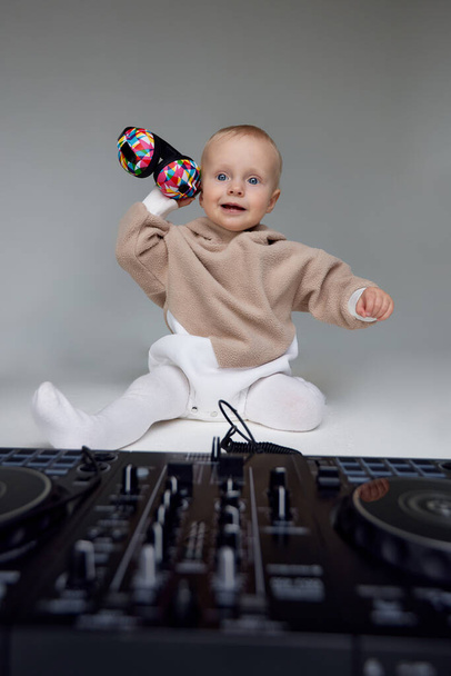 Egy aranyos mosolygós kisgyerek bézs kapucnis pulcsiban ül a padlón DJ fejhallgatóval és DJ keverőpulttal. Zene és szórakozás. Szürke háttérrel elszigetelve - Fotó, kép