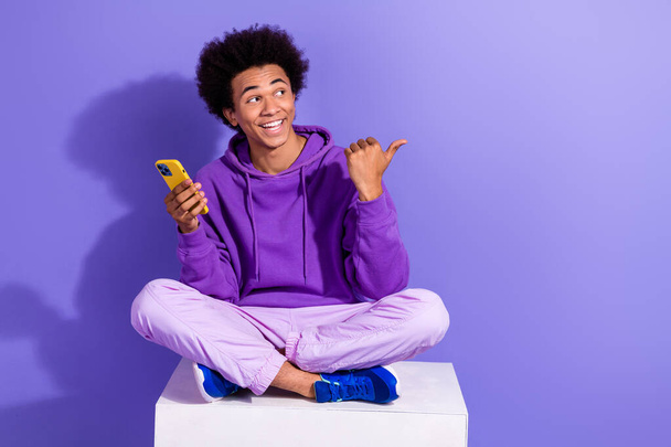 Full length photo of cheerful guy sit on white platform look directing empty space hold smartphone isolated on violet color background. - Foto, Imagen