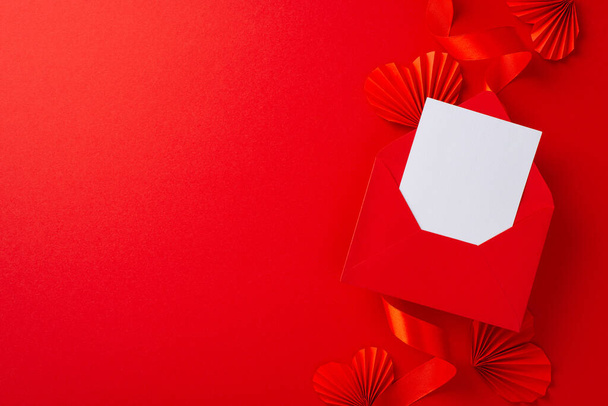 Parlak kırmızı kurdele ve kağıttan origami kalplerle çevrili beyaz boş zarfın üst görünüm fotoğrafı kopyalanmış kırmızı arka planda - Fotoğraf, Görsel