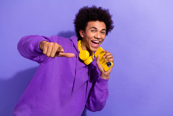 Portrait of pleasant handsome guy wear purple pullover headphones sing in smartphone directing at you isolated on violet color background. - Φωτογραφία, εικόνα