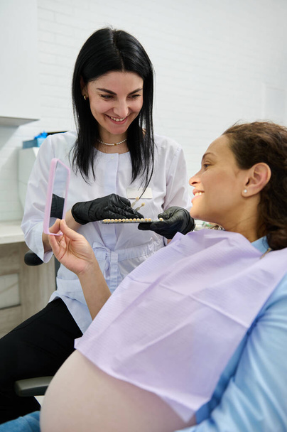 Όμορφη έγκυος γυναίκα, ασθενής σε ραντεβού με οδοντίατρο, κάθεται σε καρέκλα οδοντιάτρων, χαμογελά στον καθρέφτη, συζητώντας με το γιατρό τη σκιά και το χρώμα των δοντιών για λεύκανση διαδικασία στην οδοντιατρική κλινική - Φωτογραφία, εικόνα