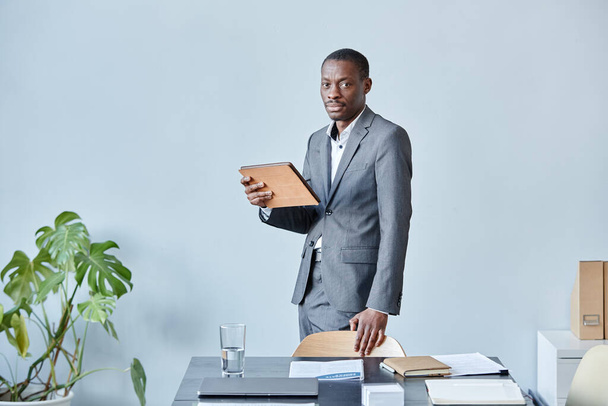 Retrato mínimo de ejecutivo negro profesional mirando a la cámara mientras está de pie contra la pared azul en la oficina con traje elegante, espacio de copia - Foto, Imagen