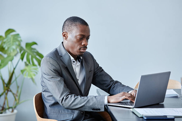 Minimalny portret profesjonalnego czarnego wykonawcy korzystającego z laptopa siedzącego w miejscu pracy przy niebieskiej ścianie w biurze i noszącego garnitur, przestrzeń do kopiowania - Zdjęcie, obraz