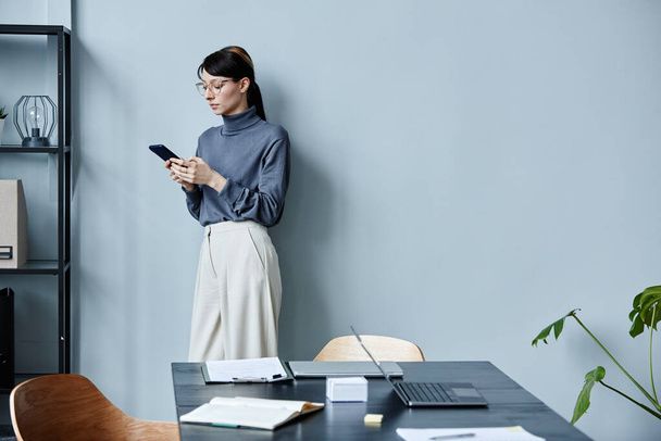 Minimales Porträt einer jungen Geschäftsfrau mit Smartphone an blassblaue Wand gelehnt und in lässigem Outfit im Büro, Kopierraum - Foto, Bild