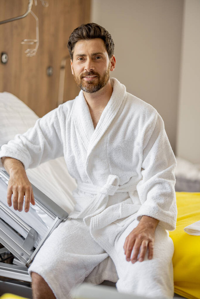 Portret van een vrolijke volwassen man die op bed zit in een afdeling van een modern ziekenhuis, rustend tijdens de revalidatie na medische behandeling - Foto, afbeelding