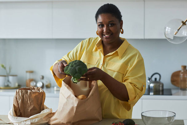 Porträt einer afroamerikanischen Hausfrau, die in die Kamera lächelt, während sie in der Küche stehende Produkte aus Papiertüten herausnimmt - Foto, Bild