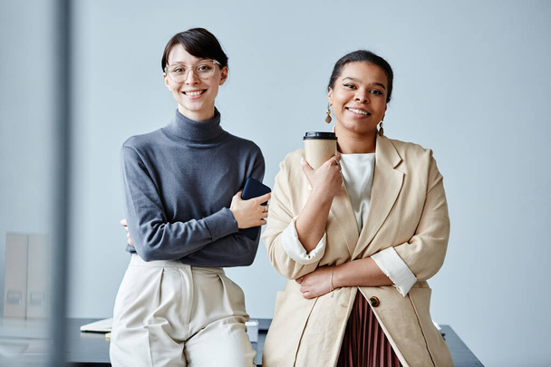 Candid waist up portrait of two young businesswomen smiling at camera in office against simple background - 写真・画像