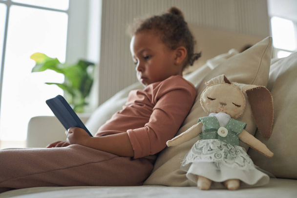 Niña afroamericana viendo dibujos animados en la tableta PC mientras está sentado en el sofá en la habitación - Foto, Imagen