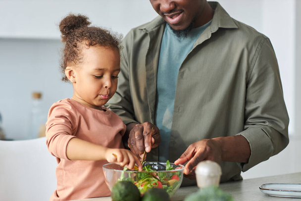 African American little girl helping her dad to cook vegetable salad in the kitchen - Photo, Image