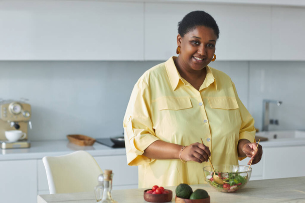 Portret van Afro-Amerikaanse huisvrouw glimlachend op camera tijdens het bereiden van groentesalade in de keuken - Foto, afbeelding