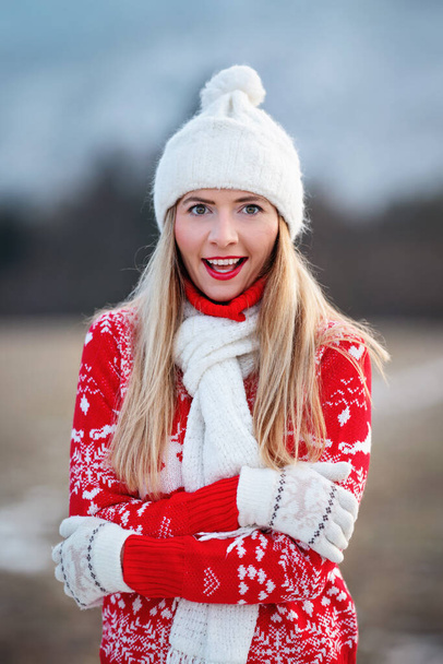 Young woman in red winter pullover, holding her elbows looking surprised, blurred country background - Photo, Image