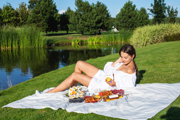 Νεαρή χαμογελαστή γυναίκα που χαλαρώνει έξω και κάνει πικ-νικ, κάθεται πάνω σε μια κουβέρτα στο γρασίδι. - Φωτογραφία, εικόνα