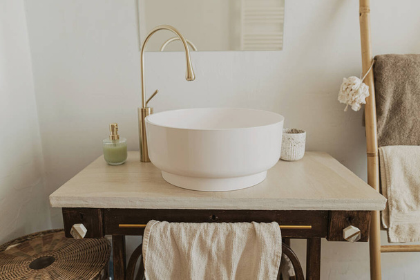 Modern ceramic sink with golden faucet on rustic stand with soap and towel on background of wall with mirror and travertine tile, modern bathroom interior. Stylish boho bathroom design. - 写真・画像
