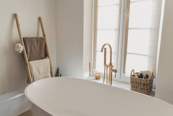 Стильний дизайн ванної кімнати. Сучасна ванна з золотим краном від підлоги, дерев'яні сходи з рушниками та великим вікном, сучасний інтер'єр еко-санвузла
. - Фото, зображення