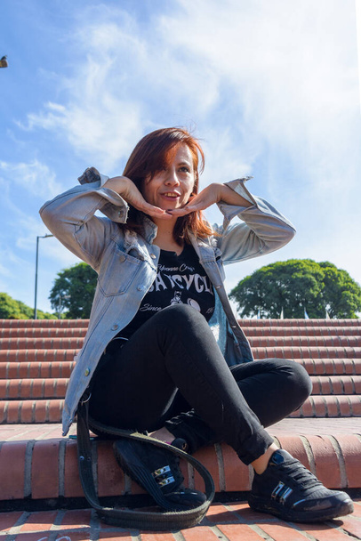 комический портрет латинской женщины колумбийской национальности с рыжими волосами и руками на подбородке, улыбающейся, сидящей в парке. Она вертикально, скрестила ноги, сидя лицом к камере. вертикальная. - Фото, изображение