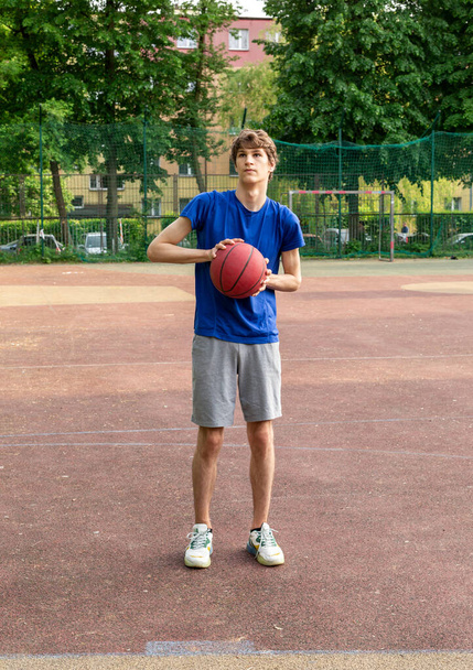 Söpö poika t-paidassa pelaa koripalloa kaupungin leikkikentällä. Aktiivinen teini nauttia ulkona peli punainen pallo. Harrastus, aktiivinen elämäntapa, urheilu lapsille. - Valokuva, kuva