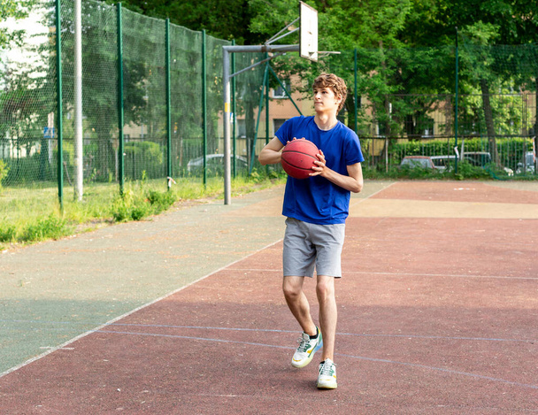 Cute boy in t shirt plays basketball on city playground. Active teen enjoying outdoor game with red ball. Hobby, active lifestyle, sport for kids. - Foto, immagini